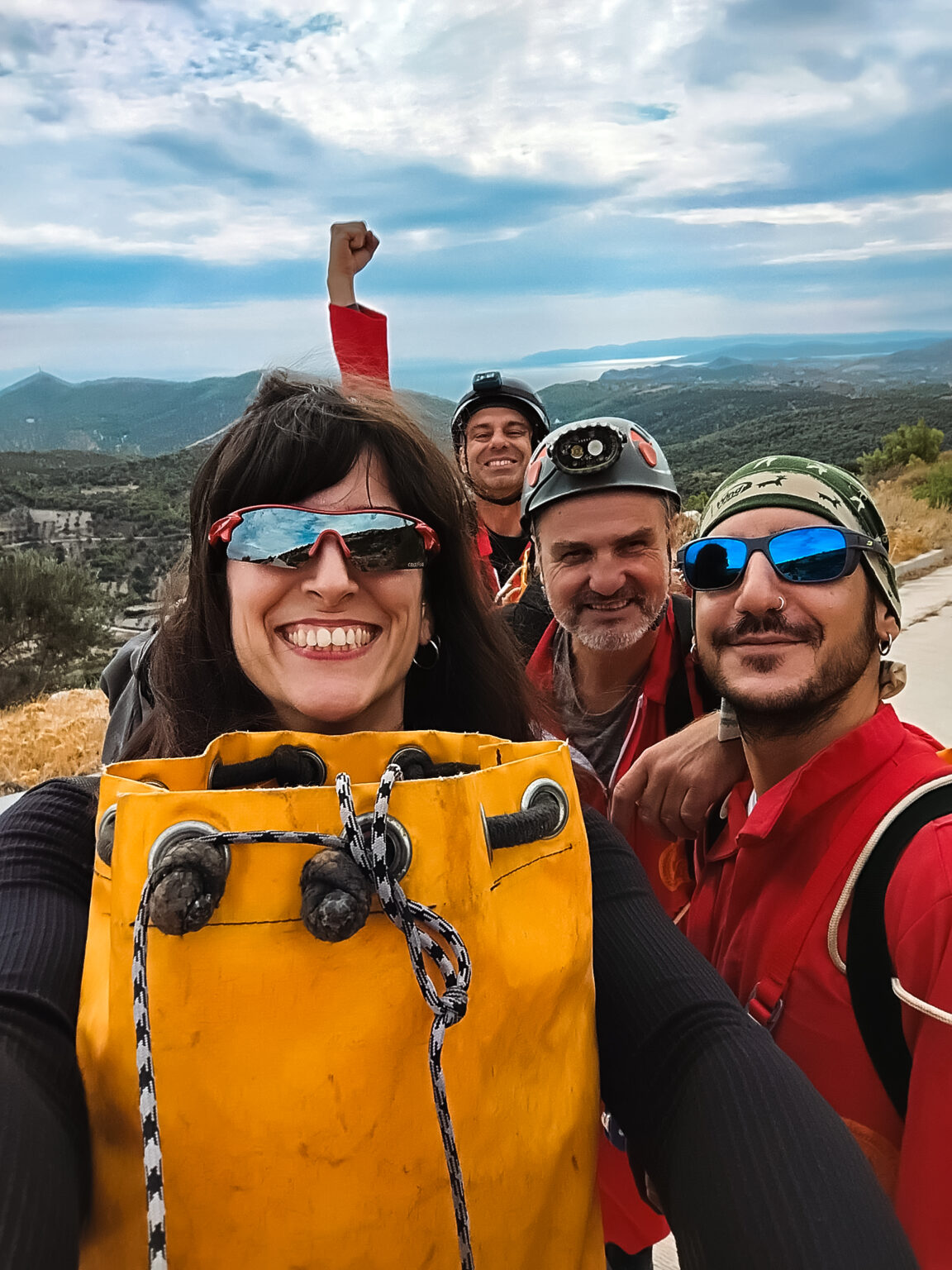 Group selfie of four smiling cavers outdoors on Mount Merenta wearing helmet, sunglasses, and caving gear, holding a yellow tackle bag.