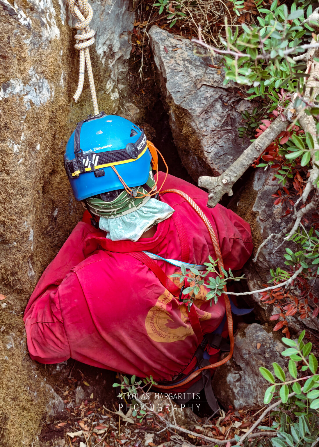 Caver wearing a blue helmet and red suit squeezing into a narrow rock fissure at the entrance of Fteri cave.