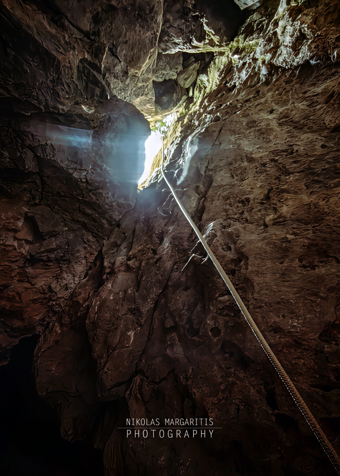 Looking up from inside a dark vertical cave shaft along a hanging rope toward a bright natural entrance opening.