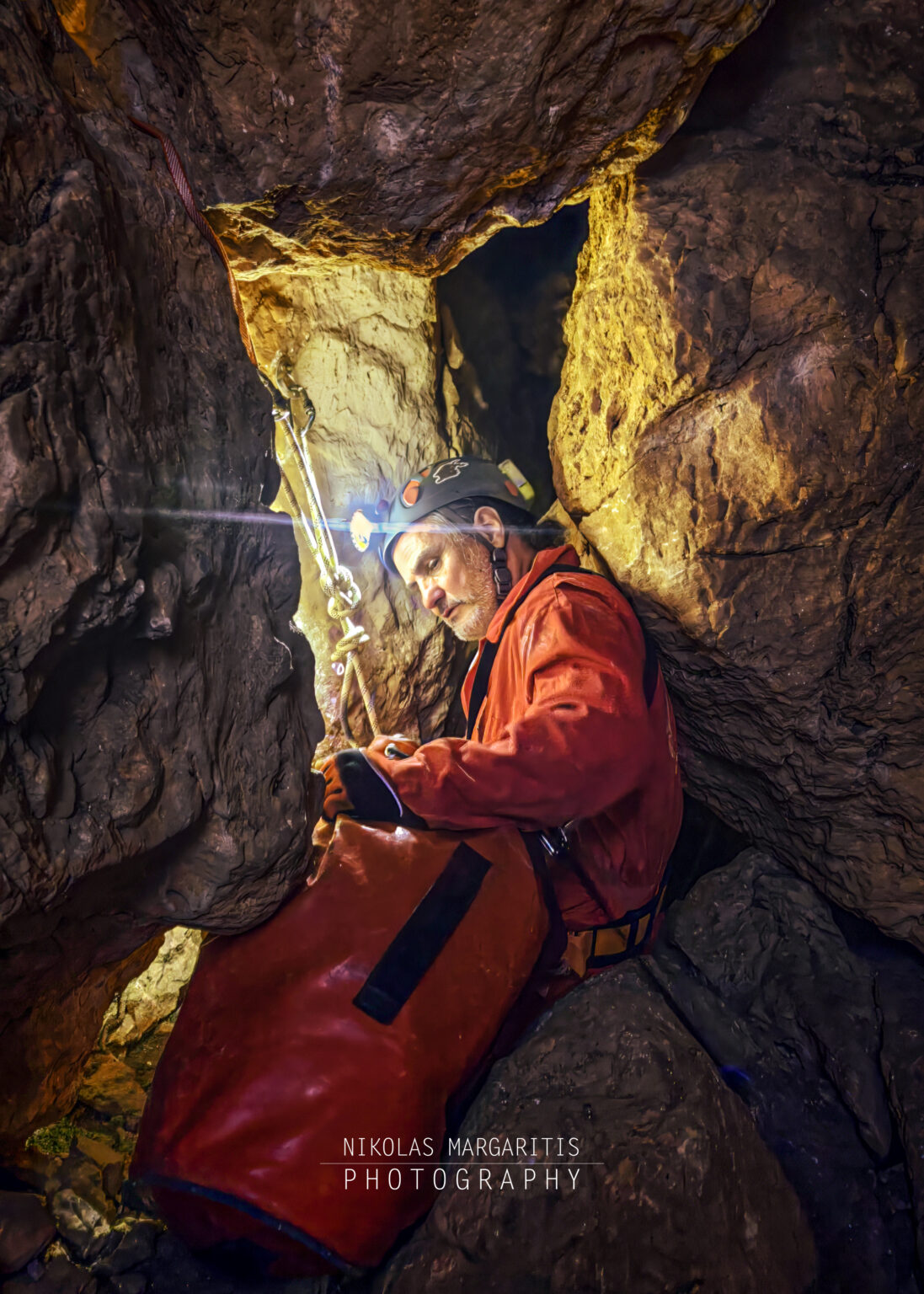 Caver in a red undersuit and helmet with a lit headlamp sitting inside a tight rocky cave passage at -10m depth, handling a red tackle bag next to a rigged rope.