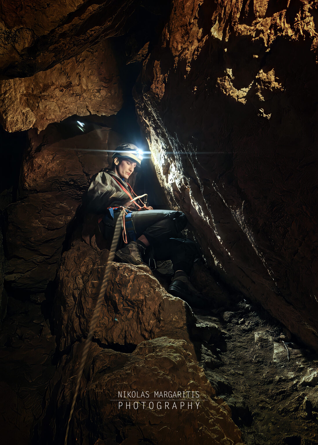 Looking down or along a vertical cave shaft at a caver descending on a rope with a headlamp lit during the P19 pitch descent in Fteri cave.