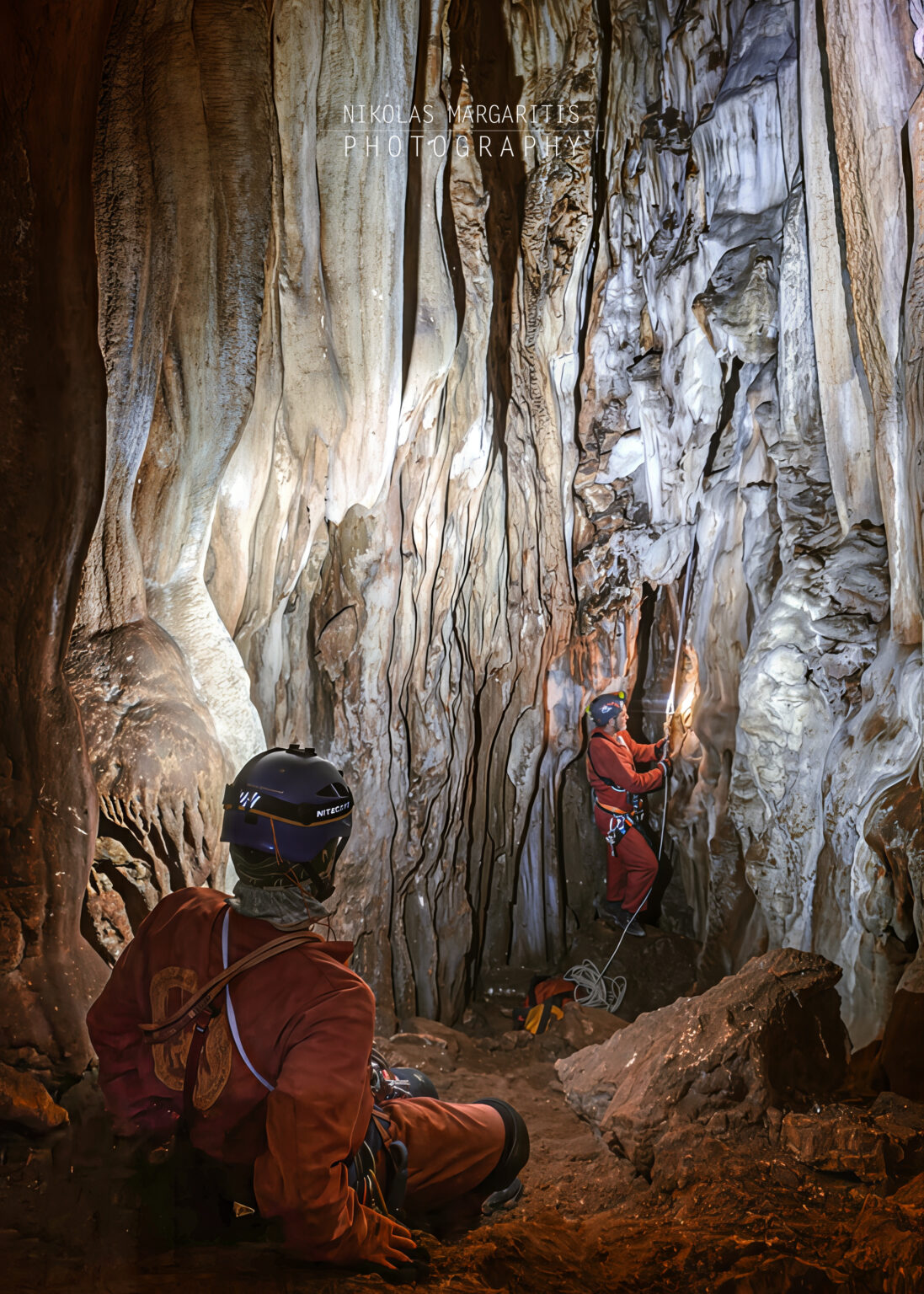 Inside a narrow cave shaft with rich flowstone formations, one caver sits in the foreground watching another caver in a red suit rig a rope near large stalactites.