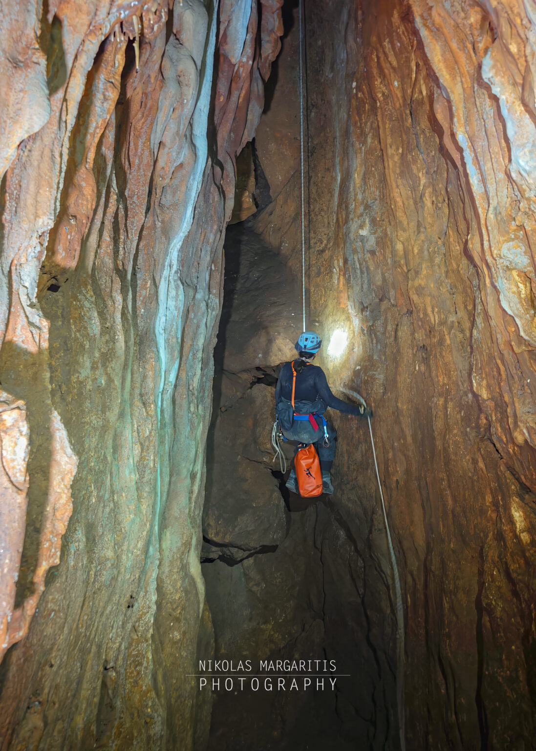 Full-length view of a caver wearing a blue helmet and dark suit descending a rope in a wide, vertical rock shaft with colorful flowstone walls and an orange tackle bag hanging below.