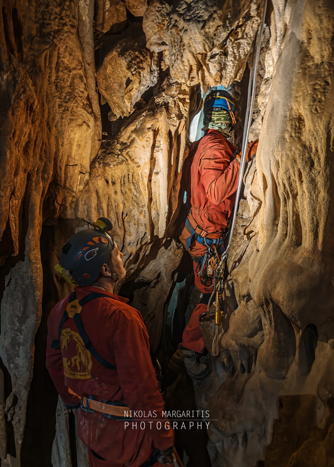 Sokratis in a red suit and helmet watching from below as Spyros squeezes through a narrow vertical rock passage lined with flowstone formations.