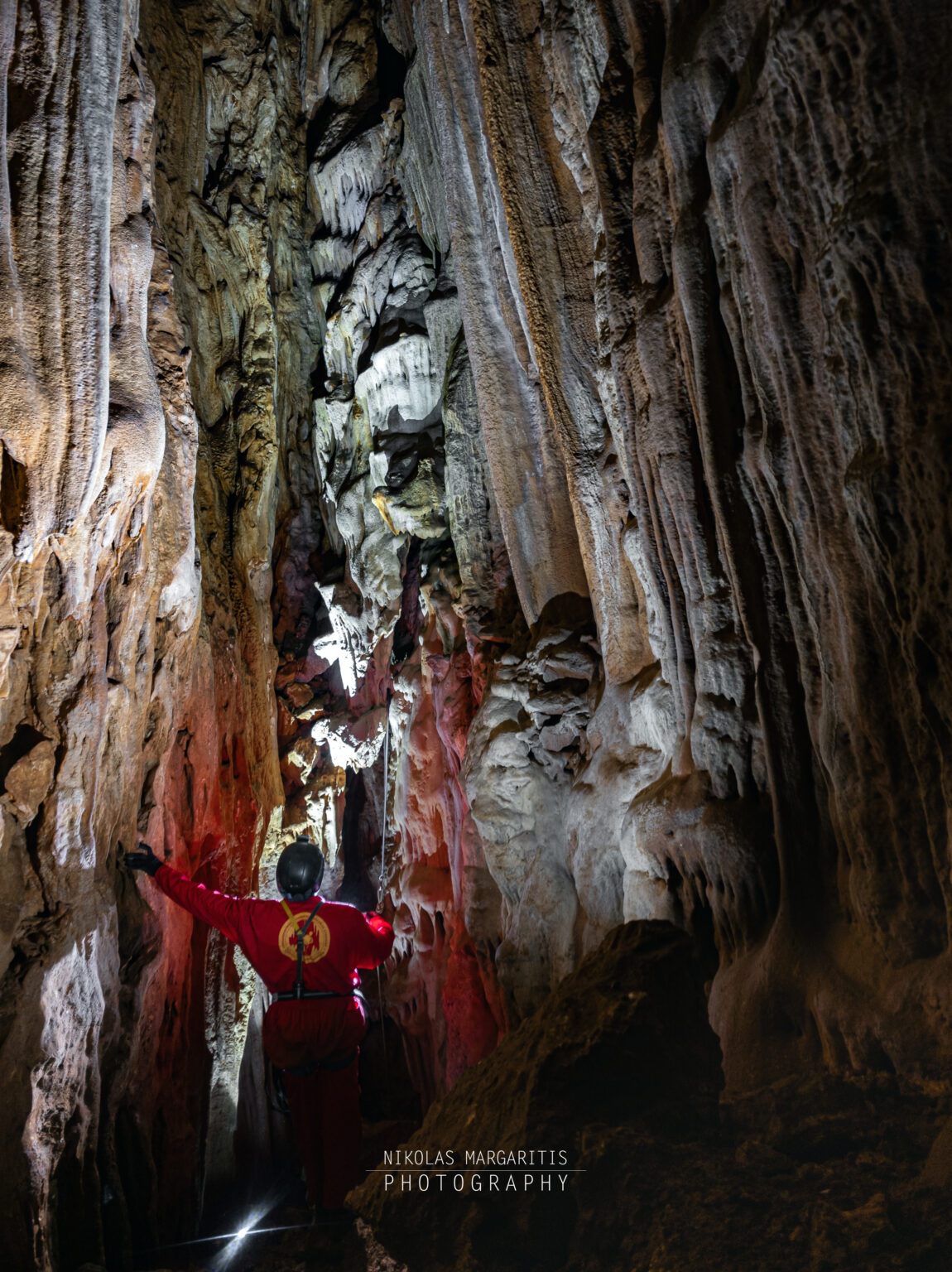 Self-portrait of caver Nikolas Margaritis in a red suit facing into a tall, narrow shaft lined with impressive stalactites and flowstone formations at -24m.
