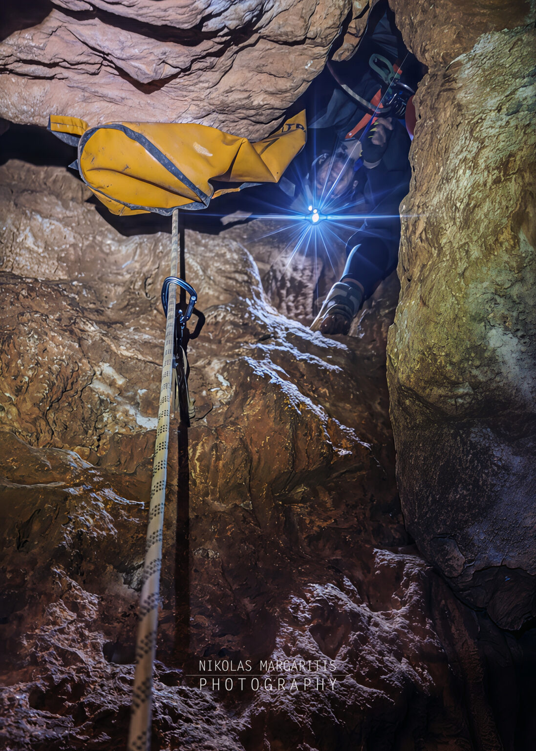 Looking up from below along a climbing rope during ascent in a vertical cave shaft, showing a caver above looking down with a brightly lit headlamp next to a yellow tackle bag.
