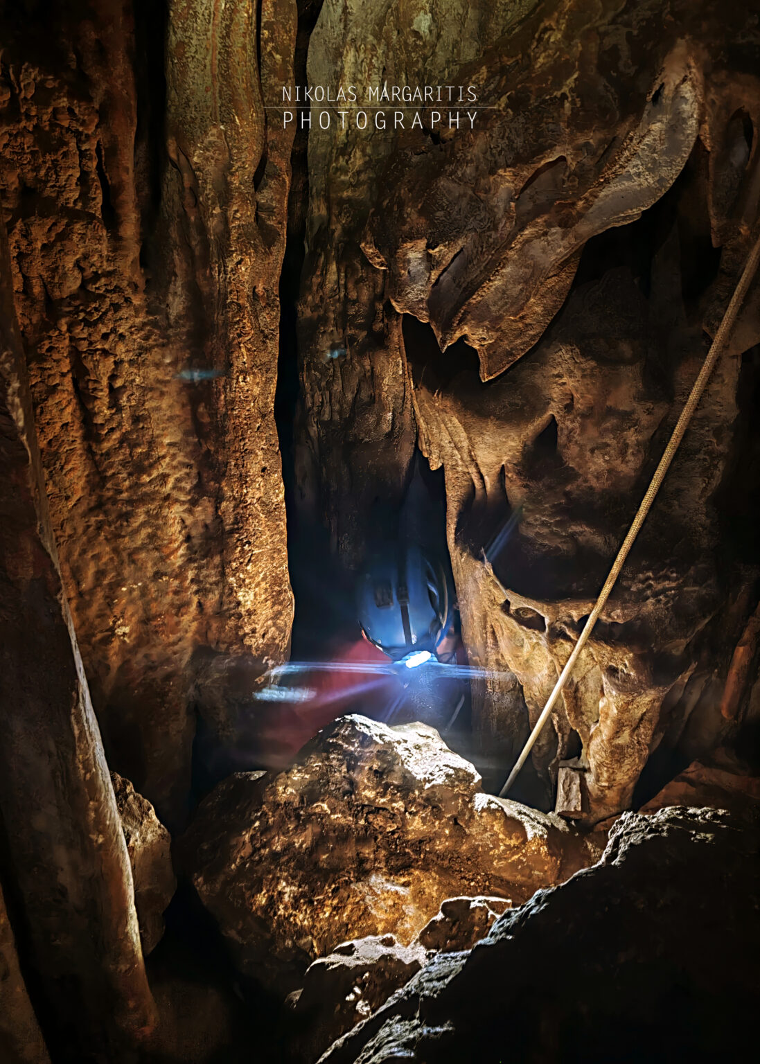 A caver wearing a blue helmet with a bright headlamp squeezing through a narrow, dark rock passage framed by carved flowstone walls.